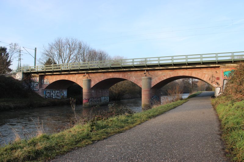 Dreifeldrige Sandstein-Eisenbahnbrücke mit drei Bögen über der Nidda in Frankfurt-Nied; daneben verläuft ein Uferweg.