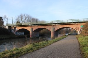 Dreifeldrige Sandstein-Eisenbahnbrücke mit drei Bögen über der Nidda in Frankfurt-Nied; daneben verläuft ein Uferweg.